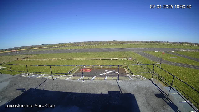 A clear blue sky fills the top half of the image, while the lower half shows a large, open grassy area with a runway. There are marked areas on the tarmac, including a red section and a symbol that resembles an airplane runway. A wooden fence lines the grassy area, and several taxiways can be seen leading off the main runway. The scene is viewed from a tower or building, indicated by a railing in the foreground. The timestamp in the corner shows the date and time as 07 April 2025 at 16:00:49.
