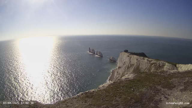 A panoramic view of the ocean under a clear blue sky, with sunlight reflecting off the water, creating a shimmering effect. In the foreground, a chalk cliff with grassy terrain leads to steep edges. Several large rock formations, resembling sails, rise from the water in the middle ground. A small building or lighthouse is located at the top of the cliff on the right side. The scene conveys a serene coastal landscape.