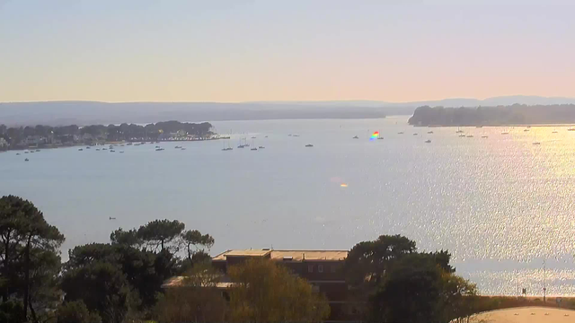 A serene view of a tranquil body of water reflects the sun, creating a sparkling surface. Various boats are scattered across the water, while trees line the shore on the left. In the distance, hills rise gently against a light-colored sky, creating a peaceful landscape. The scene conveys a calm and pleasant atmosphere.