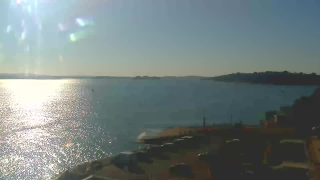 A clear sky with bright sunshine creates a shimmering reflection on a calm body of water. In the foreground, there is a parking area with several vehicles. The water extends towards the horizon, where faint outlines of distant land are visible. The scene conveys a serene and peaceful atmosphere, typical of a sunny day by the water.