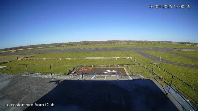 A clear blue sky fills the upper part of the image. In the foreground, there is a flat rooftop with a railing, and a section of a white structure is visible on the left. Below, an expansive green grass field stretches out, intersected by a runway marked with light and dark lines. A red windsock stands near the edge of the field, and a wooden fence borders the grassy area. The overall scene appears bright and sunny, indicating a pleasant day.