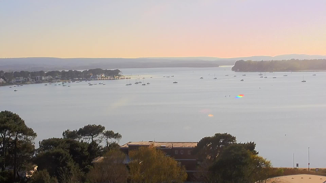 A serene water scene shows a wide bay with several small boats scattered across the surface. In the background, there are trees and buildings lining the shore. The sky transitions from soft pastel colors to a clear horizon with distant hills, creating a peaceful ambiance.