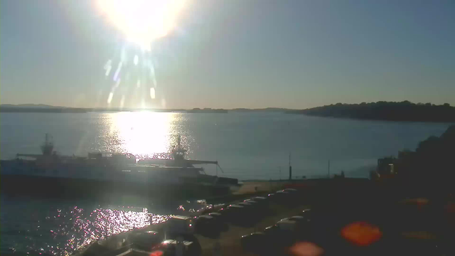 The image shows a waterfront scene during daylight. The sun is bright in the sky, casting a glare over the water, which reflects shimmering light. A large boat is docked at the shore, and several vehicles are parked nearby. In the background, a distant shoreline is visible with hills under a clear blue sky.