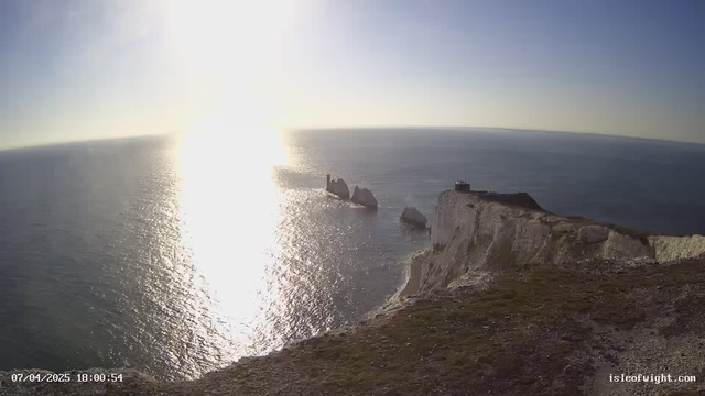 A scenic view of the ocean taken from a high vantage point. The sunlight reflects off the water, creating a shimmering effect. In the foreground, there is a grassy cliff edge leading down to the water. Visible in the distance are several large rock formations emerging from the sea. The sky is clear with a few wispy clouds. The bottom left corner shows a timestamp indicating the date and time of the image.