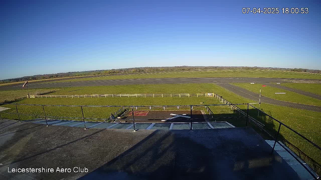 A wide view of an airfield taken from a webcam. The sky is clear and blue. In the foreground, a roof or balcony railing is visible, with a tarmac area below that shows markings for air traffic. The airfield features grassland on either side, with a light-colored fence surrounding part of the area. In the distance, there are patches of trees and a flat landscape extending to the horizon. The image shows the date and time in the top right corner.