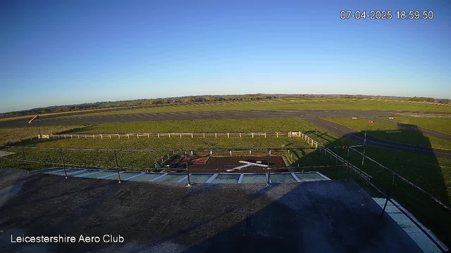 A clear blue sky stretches across the image, with a vast green landscape below. In the foreground, there is a flat rooftop with a railing, and a large, white cross marked on the ground. To the left, a windsock indicates wind direction. In the background, an airstrip runs horizontally with a grassy area adjacent to it. The scene is bright, suggesting it is daytime.