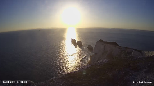 A coastal scene at sunset, showing the sun low in the sky casting golden reflections on the calm ocean. In the foreground, white cliffs rise from the water, while two rock formations jut out from the sea. The horizon is visible in the distance, with a soft gradient from blue to orange as the sun sets. The image has a serene and tranquil atmosphere.