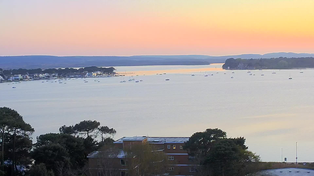 A serene view of a calm bay during sunset. The sky is a gradient of soft pastel colors, blending from light orange to pale pink and blue. In the water, several small white boats are anchored, reflecting the colorful sky. On the left, a row of buildings with varying colors stands along the shoreline, framed by trees. In the distance, gentle hills rise on the horizon, completing the peaceful landscape.