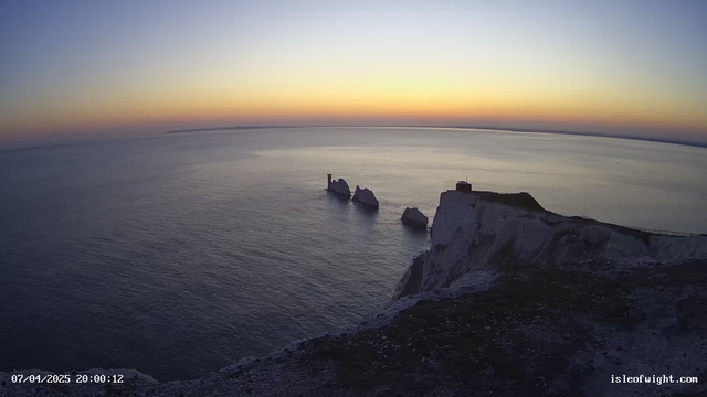 A scenic view of the ocean at sunset, with a gradient of colors in the sky ranging from orange to blue. In the foreground, a cliff with white rocks and sparse green vegetation descends towards the water. Three prominent rock formations jut out of the water, partially illuminated by the fading sunlight. A small structure is visible atop the cliff to the right. The calm sea reflects the colors of the sky. In the distance, a faint outline of land is visible on the horizon.