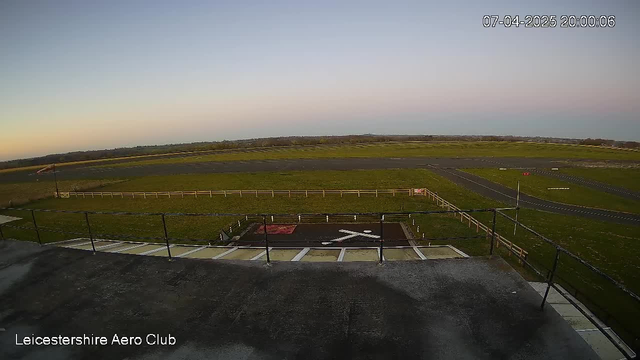 A view from a webcam positioned at the Leicestershire Aero Club, showing an open field with a grassy landscape. In the foreground, there is a fence and a tarmac area marked with a white "X." The sky is transitioning from light blue to a soft sunset hue, indicating evening time. The image conveys a peaceful and clear atmosphere, with no visible aircraft or people in the scene.