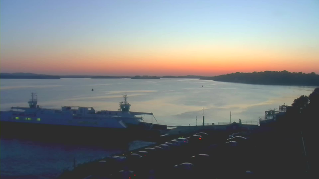 A calm body of water reflects soft shades of blue, pink, and orange as the sun sets in the background. Two ferries are docked at the shoreline, with one ship partially visible on the left side of the image. The shoreline features a few boats and a faint outline of trees and hills in the distance. A line of parked cars is visible in the foreground, adding to the serene setting.