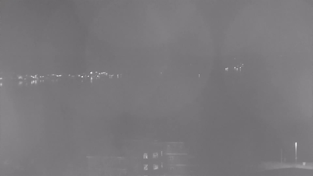 A low-contrast black and white image depicting a blurry waterfront scene at night. Faint light reflections can be seen on the water surface, with scattered points of light indicating distant buildings or street lamps. The foreground features an indistinct structure, likely a building, appearing obscured and low in detail.