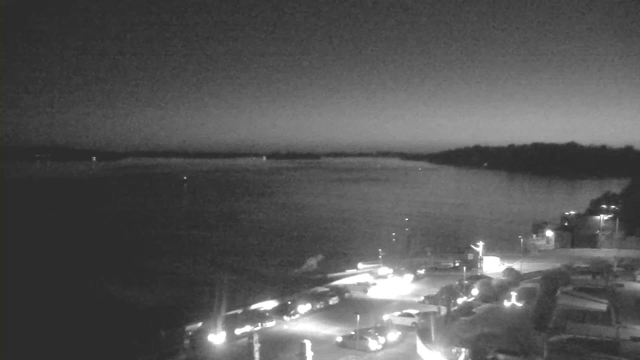 A grayscale image of a waterfront scene during twilight. The horizon is dimly illuminated, reflecting light from the water. In the foreground, there are several parked vehicles along a road, with some light sources visible, possibly from streetlights or vehicle headlights. The background shows faint outlines of trees and shoreline against a darkening sky. The overall mood is calm and serene, suggesting evening time by the water.