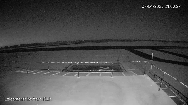 The image shows a dimly lit scene from a webcam overlooking an airfield at night. There is a dark runway visible in the foreground, marked with white lines. A small fenced area is situated centrally, with an open space around it. The background features a horizon with faint lights, indicating a distant landscape. The overall ambiance is nighttime with low visibility, creating a quiet, tranquil setting. The bottom corner displays the text "Leicestershire Aero Club" along with a timestamp.
