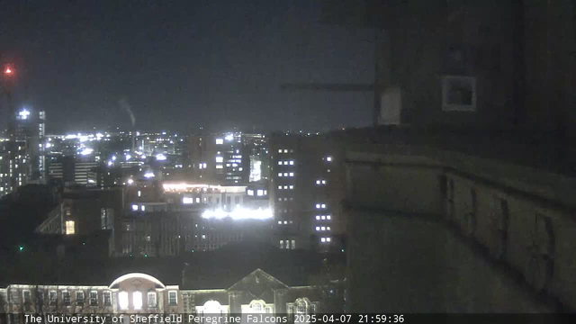 A nighttime cityscape view from a webcam at the University of Sheffield. In the foreground, dark architectural features are visible, while the background showcases a city filled with lights from various buildings. Some of the buildings have illuminated windows, creating a contrast against the darker sky. A few clouds can be seen, and there is a red light in the upper left corner, possibly from a communication tower. The overall scene conveys a calm urban atmosphere at night.