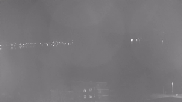 A dimly lit scene captured by a webcam at night. In the foreground, there is a blurred, dark area that appears to be a building with light reflecting off its windows. In the background, a faint outline of water is visible with small lights reflecting on the surface, likely from distant boats or shorelines. The overall image has a grainy quality.