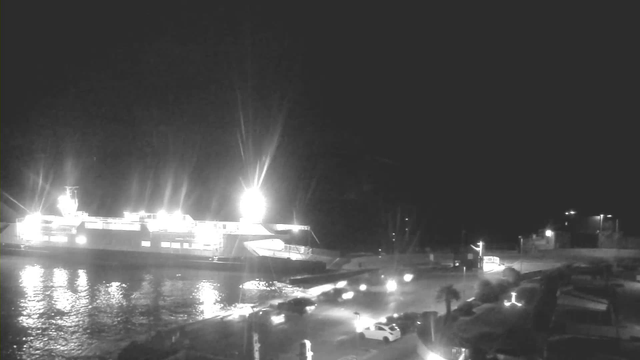A nighttime image of a harbor with a lit ferry docked on the left side. The ferry is bright with several lights illuminating its structure and the water around it. In the foreground, there are parked cars and a dark road leading away from the harbor. The water reflects the lights but appears mostly dark, and there are some trees and structures visible in the background. The overall scene is dimly lit, creating a tranquil atmosphere.