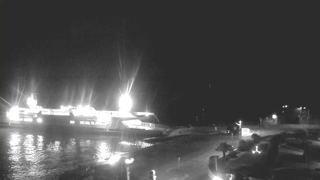 A dimly lit scene shows a large ship docked at a pier, illuminated by bright lights on the vessel. The water reflects these lights slightly, creating a shimmering effect. In the foreground, there are structures and equipment that appear to be part of the docking facility, including parked vehicles and poles. The area is surrounded by darkness, with distant lights visible along the shoreline, indicating structures or pathways.