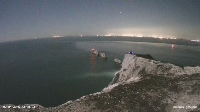 A night view of a coastline with rocky cliffs and the ocean. The water is calm, reflecting faint lights from distant land. Two large rocks protrude from the water. There are small lights on one of the rocks, indicating possible activity or a structure. The sky is starry with a faint haze, and subtle city lights are visible along the horizon. The scene conveys a tranquil, nighttime atmosphere.
