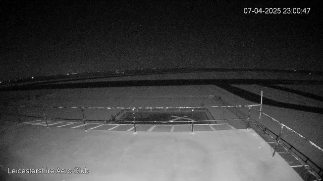 A dark image taken from a webcam at Leicestershire Aero Club. The scene features a flat rooftop with a railing in the foreground. In the background, there is an open area, possibly an airfield, with faint lights visible in the distance. The image is predominantly black, suggesting it was captured at night. The timestamp indicates the date and time of the capture.