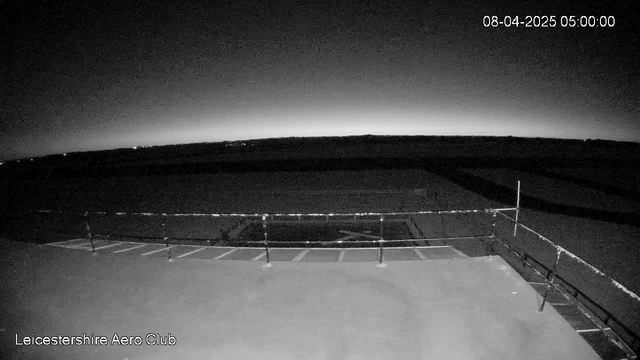 A dark, panoramic view of a landscape at dawn, with a faint gradient of light on the horizon. In the foreground, there is a railing along a flat surface, suggesting a viewing platform or rooftop. The edge of the platform features a grid pattern that leads towards an open space, indicating the presence of an airfield or runway in the distance. The timestamp in the top right corner shows the date and time as April 8, 2025, at 5:00 AM.