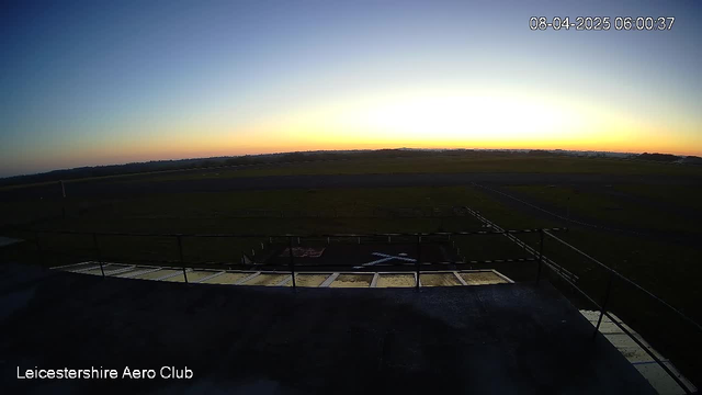 A panoramic view of a clear sky at dawn, showing a gradient of colors from deep blue at the top to warm orange near the horizon. Below the sky, there is an expanse of green grass, likely part of an airfield, with a concrete platform in the foreground. An airplane outline is faintly visible on the ground. The time and date in the top right corner indicate 06:00:37 on August 4, 2025.