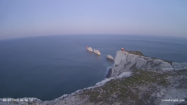 A coastal view showing rocky cliffs that meet the blue sea. In the foreground, there is a grassy area with white stones. In the distance, several large white rocks emerge from the water, with a red lighthouse on one of them. The sky is light blue with soft clouds, indicating early morning light.