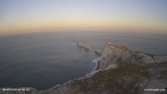 A panoramic view of the coastline during dawn, featuring rugged white cliffs on the right and the shimmering blue sea extending into the horizon. The sky transitions from soft orange at the horizon to a pale blue overhead. In the distance, there are two large white rocks emerging from the water, with a red lighthouse situated on one of them. To the right, a building is perched on the cliff, illuminated by early morning light.