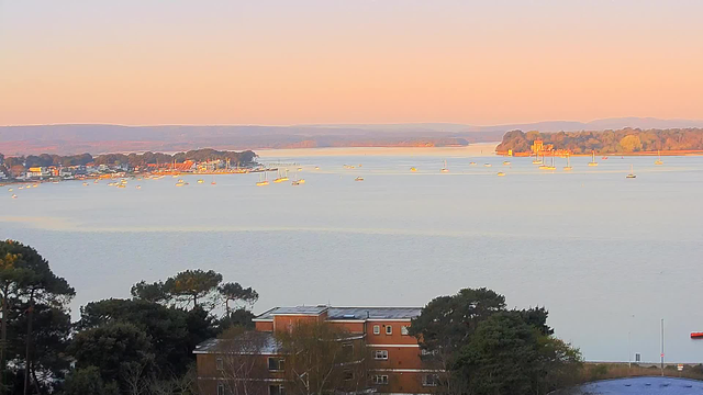 A serene waterfront view during sunset, showcasing a calm expanse of water scattered with small white boats. The shoreline is lined with trees and buildings, with a mix of green and orange hues reflecting the warm colors of the sunset. In the distance, low hills are faintly visible against a soft gradient sky transitioning from peach to lavender.