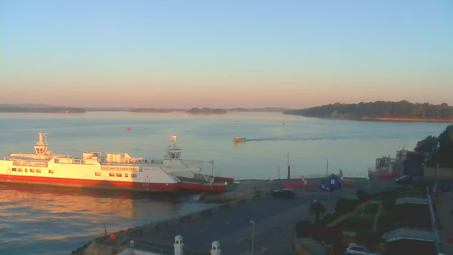 A view of a calm waterway during early morning light. In the foreground, a large white ferry with red accents is docked at the shore, with the words "GIVE WAY TO CHAIN FERRY" visible on its side. A small yellow boat is navigating in the water nearby. The water reflects the soft colors of dawn, while a few buoys and distant land are visible across the calm surface. The shoreline is lined with trees, and there are some buildings and vehicles near the water's edge, framed by the softness of the morning sun.
