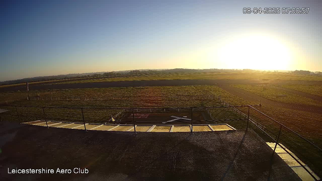 A wide view of a grassy airfield at sunrise. The sky is clear with a gradient from deep blue at the top to a warm orange near the horizon. A small section of the pavement is visible in the foreground, leading to the airfield, which has a simple white aircraft marking on the ground. Fencing lines the edge of the field, and a few distant rolling hills are visible in the background. The atmosphere is tranquil, indicating the early morning time. The lower left corner displays the words "Leicestershire Aero Club" in white text.