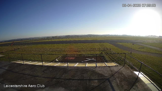 A wide view of a grassy airfield under a clear blue sky. In the foreground, there are stairs leading down from a raised platform, with a railing around it. On the ground, a red and white windsock is positioned to the left, and on the airfield, there are markings including a large white "X". The background features distant trees and hills under bright sunlight. The image is timestamped with the date and time in the upper right corner.