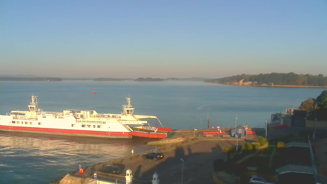 A white and red ferry is docked at a harbor with calm blue waters surrounding it. In the background, a distant shoreline with trees and rocky cliffs can be seen under a clear blue sky. The foreground shows a pavement area with a few parked cars and structures, while some people are walking near the dock. A buoy is visible floating in the water.