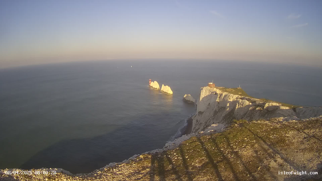 A panoramic view of a coastal landscape under a clear sky. The foreground shows rocky cliffs with patches of grass and scattered stones. In the distance, there are three white chalk cliffs rising from the water, with two lighthouses visible on them. The ocean is calm, reflecting the blue and soft orange hues of the sky, suggesting early morning light. The horizon line is clear, and a few distant boats are visible on the water. Shadows extend from the foreground, indicating the position of the sun.