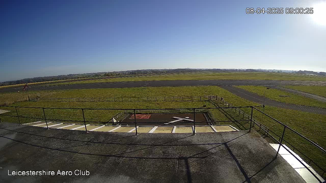 A clear blue sky spans the upper portion of the image, with the sun bright in the top right corner. Below, there is a green field stretching across the midsection, bordered by a paved runway on the right and a helipad marked with a white cross on the left. Fencing outlines the area, and a small structure is visible in the foreground. The setting appears to be an open-air space near an aerodrome, with no aircraft present in view. The time stamp in the corner indicates it is 09:00 on April 8, 2025.