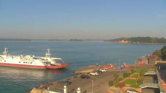 The image shows a calm body of water with a ferry docked at a terminal on the left side. The ferry is white with red accents and has vehicles on board. In the foreground, there is a parking lot with several cars. To the right, there are some trees and a small building near the water. The sky is clear and blue, and the landscape in the distance features a coastline with greenery and cliffs.