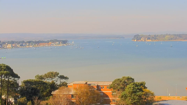 A panoramic view of a calm body of water, with several small boats sailing. The shoreline features a small town with buildings, and trees line the waterfront. In the background, rolling hills can be seen under a clear sky. The scene is bathed in soft sunlight, creating a tranquil atmosphere.