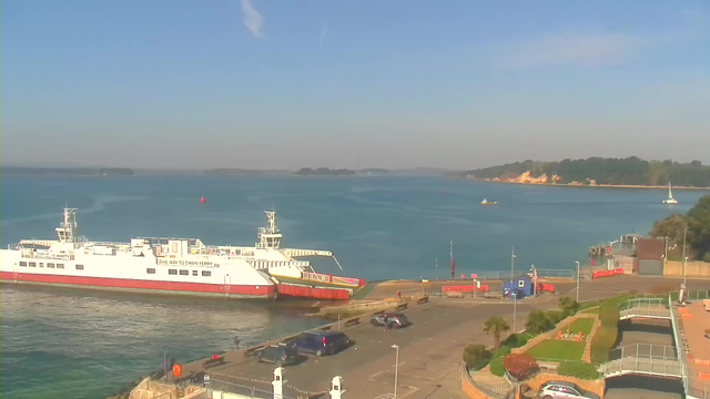 A sunny coastal scene featuring a large white ferry docked at a terminal with red accents. The water is calm and blue, with several boats in the distance. A parking area is visible near the dock, with a few cars parked. In the background, there are green trees and a shoreline with sandy cliffs. The sky is clear with a few wispy clouds.