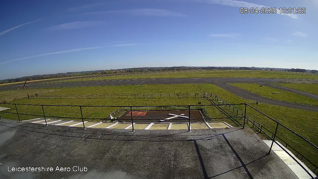 A wide view from a webcam positioned on a rooftop overlooking an airport. The image features a clear blue sky with a few wispy clouds. Below, there is a large grassy area that leads to a runway, marked with a few taxi lines. In the foreground, a fenced area contains a red square and a white cross on the ground. The airport is mostly empty with minimal activity visible. The scene is bright and clear, indicating a sunny day.