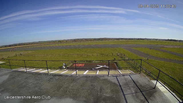 A clear daytime view of an aerial landscape featuring a grassy airstrip. There is a small distinct "X" marking on the ground near the center, indicating a landing area. A balcony railing is visible in the foreground, and a lone person is seated at the edge of the airstrip. The sky is bright blue with some faint white clouds, and the surrounding land is mostly flat and green, with a few distant hills in the background. The scene conveys a sense of calm and open space.