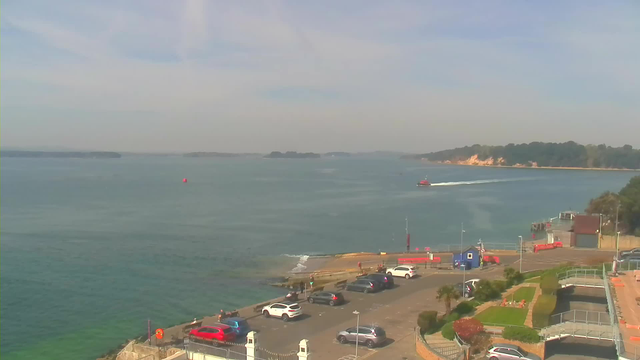 A coastal view showing a calm sea with a small red boat moving through the water. In the foreground, a parking area is lined with various vehicles including cars and SUVs. A walkway with green grass borders the water, and a few trees can be seen in the background. The sky is clear with some light clouds. On the horizon, small islands are visible amidst the water.