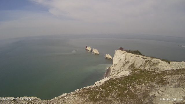 A scenic view overlooking a calm sea with a hazy sky. In the distance, white chalk cliffs rise above the water. Three prominent rock formations, similar in color to the cliffs, jut out from the sea. A red and white striped lighthouse is visible on one of the rocks. A small boat is traveling in the water near the formations. The foreground features grass and rocky terrain, blending into the cliffs.