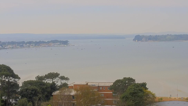 A wide view of a calm body of water, likely a bay, with several small boats scattered on the surface. In the distance, a shoreline with houses and trees is visible, along with a breakwater and some structures. The sky is overcast, creating a muted color palette. In the foreground, greenery and a low-rise brick building can be seen. The overall atmosphere is peaceful and serene.