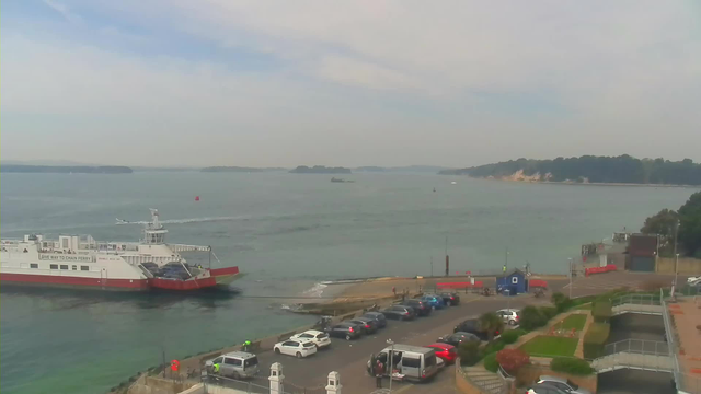 A calm water scene featuring a chain ferry moving across the water, with a parking area visible in the foreground. Several parked cars are arranged along the waterfront, and there are a few people walking near the ferry terminal. In the distance, small boats can be seen on the water, alongside a tree-lined shoreline. The sky is mostly cloudy with hints of blue peeking through.