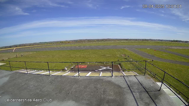 A wide view of an airport landscape under a clear blue sky with a few clouds. In the foreground, there is a railing and part of a rooftop; the ground below has green grass and some light-colored markings. In the distance, there are multiple runways and a few small planes visible. A windsock stands to the side, indicating the direction of the wind.