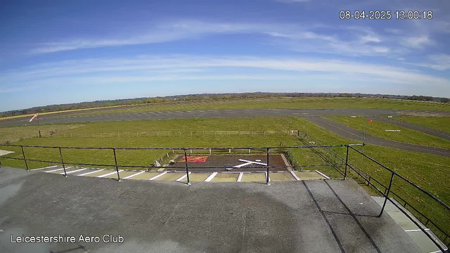 A view from a webcam at Leicestershire Aero Club shows a clear, sunny sky with a few scattered clouds. In the foreground, there is a railing along the edge of a flat rooftop. Below, an airstrip extends into the distance, flanked by green grass and sections of gravel. A windsock is visible, indicating the wind direction. The surroundings are mostly open fields with a few trees in the background. The timestamp in the corner reads 13:00 on April 8, 2025.