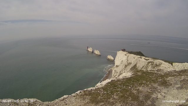 A coastal scene featuring white chalk cliffs rising sharply from the sea. In the background, the water is calm and exhibits various shades of blue and green. There are several large, jagged rocks jutting out of the water, with two small structures visible atop them. The sky is overcast with grey clouds, creating a muted atmosphere. The foreground shows grassy terrain with some rocky areas. The image includes a timestamp in the bottom left corner indicating the date and time.