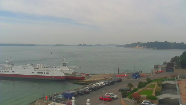A view of a calm body of water with a ferry docked at a terminal on the left. Several parked cars are visible along the waterfront, with a parking lot filled with a variety of vehicles. In the background, there are lush green hills and a distant landmass. The sky is overcast with a few clouds. On the right, there is a small building and some green landscaping near the water's edge.