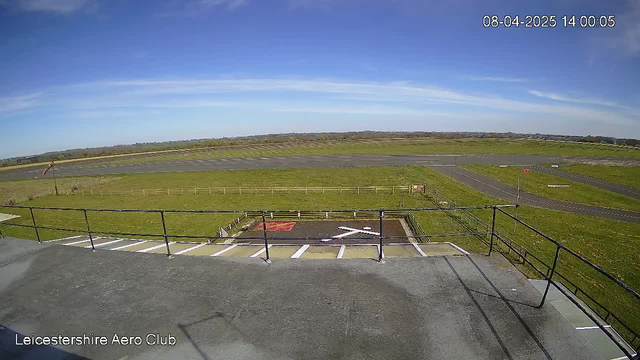 A clear blue sky with some wispy clouds stretches above a grassy landscape. In the foreground, there is a flat rooftop railing. Below, a tarmac runway is visible, lined by green grass and a wooden fence. An area on the ground features a red and white cross symbol. A red windsock is situated to the left, indicating wind direction. The scene is tranquil and well-lit, suggesting a sunny day at an airfield.