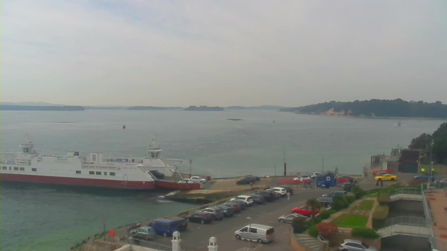 A view of a calm harbor with a large white and red ferry docked at a terminal. Several cars are parked along a road beside the water. In the background, a distant shoreline with trees and a few small boats can be seen on the water. The sky is mostly cloudy, giving a grayish appearance. The scene conveys a serene maritime atmosphere.